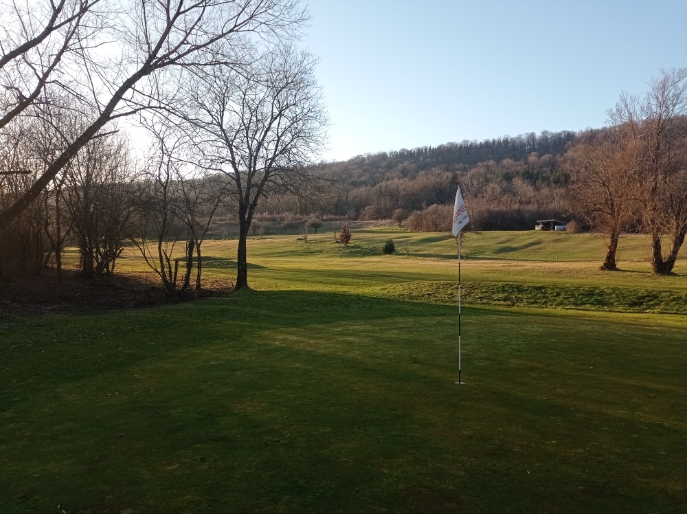 Vue panoramique d'un parcours de golf au crépuscule, mettant en avant un green avec un drapeau blanc. En arrière-plan, des collines boisées et des arbres dénudés créent une atmosphère paisible. Le sol est recouvert d'herbe verte bien entretenue, tandis que le ciel est dégagé, illuminé par la lumière douce du soleil couchant. Cette image évoque la tranquillité et la beauté d'une journée de golf en pleine nature.
