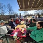 Groupe d'amis profitant d'un moment convivial en extérieur autour d'une table, avec des boissons et des collations. L'environnement est ensoleillé, entouré d'arbres, et les participants semblent se détendre après une activité de golf. Cette image illustre des retrouvailles amicales, parfaites pour promouvoir des événements sportifs ou des sorties en groupe.