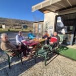 Groupe d'amis profitant d'un moment convivial en extérieur, assis autour d'une table colorée sur une terrasse ensoleillée. En arrière-plan, un paysage naturel avec des collines verdoyantes. Les participants, habillés de manière décontractée, semblent apprécier des boissons et des collations, créant une ambiance chaleureuse et amicale. Ce cadre est idéal pour des activités de loisirs, comme le golf, visible sur la gauche de l'image.