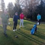 Quatre golfeurs se préparent à jouer sur un parcours de golf ensoleillé. Ils sont vêtus de vêtements de sport adaptés, avec des sacs de golf à leurs côtés. L'un d'eux s'apprête à frapper la balle, tandis que les autres observent. En arrière-plan, on aperçoit des arbres et un panneau d'information sur le parcours. Cette scène illustre une journée typique de golf, mettant en avant l'esprit de camaraderie et le plaisir du