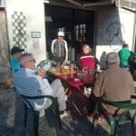 Un groupe d'hommes se détend sur une terrasse ensoleillée, devant un bâtiment d'une association sportive. Ils sont assis autour de tables colorées, profitant de boissons rafraîchissantes après une activité sportive. L'ambiance est conviviale, avec des échanges animés entre les participants. En arrière-plan, on aperçoit un panneau indiquant le nom de l'association, tandis que des éléments de la nature environnante ajoutent à la sérénité du lieu. Cette