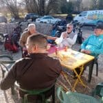 Un groupe d'hommes se retrouve autour d'une table en extérieur, partageant un moment convivial après une partie de golf. Ils sont assis sur des chaises en plastique, avec des clubs de golf visibles en arrière-plan. Des boissons sont posées sur la table, créant une ambiance détendue. L'arrière-plan montre des voitures garées et un paysage naturel, suggérant un cadre agréable pour les amateurs de golf. Cette image illustre parfaitement l'esprit de camaraderie et de lois