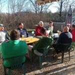 Un groupe d'amis se réunit autour d'une table en extérieur, profitant d'une journée ensoleillée. Ils discutent et partagent des boissons dans un cadre naturel, entourés d'arbres dénudés. L'ambiance est conviviale, reflétant des moments de détente et de camaraderie. Les participants sont habillés de manière décontractée, adaptés à la saison. Cette scène illustre des échanges amicaux et des instants de bonheur partagés en plein
