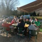 Groupe d'hommes réunis autour d'une table en extérieur, profitant d'un moment convivial en plein air. Ils dégustent des boissons, probablement des bières, et partagent des rires sous un soleil éclatant. L'ambiance est décontractée, avec des chaises en plastique et un cadre naturel. En arrière-plan, on aperçoit des voitures stationnées et des montagnes, ajoutant à l'atmosphère paisible de cette rencontre amicale.