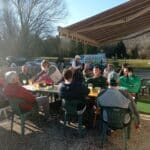 Groupe de personnes assises autour d'une table en plein air, profitant d'un moment convivial. L'image montre des hommes et une femme partageant des boissons et des plats, dans un cadre ensoleillé. En arrière-plan, on aperçoit des voitures et des participants à une activité de plein air, suggérant une ambiance détendue et amicale. Cette scène évoque des rencontres sociales, idéales pour des sorties en groupe ou des événements en extérieur.