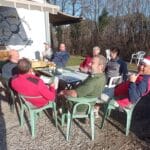 Un groupe d'hommes et de femmes se réunit autour d'une table en extérieur, profitant d'une journée ensoleillée. Ils sont assis sur des chaises en plastique, discutant et partageant des boissons et des collations. L'ambiance est conviviale, illustrant un moment de détente après une activité sportive, probablement sur un terrain de golf, comme le suggère le logo de l'Association Sportive visible en arrière-plan. Cette image reflète la camaraderie et