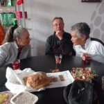 Groupe de personnes discutant autour d'une table, avec des boissons et des collations. Sur la table, on peut voir un pain, des bonbons et un plat, ce qui suggère un moment convivial. L'arrière-plan présente des vêtements de sport accrochés, indiquant un environnement lié à l'activité physique ou à un club sportif. Cette image capture l'esprit de camaraderie et de partage lors d'une rencontre informelle.