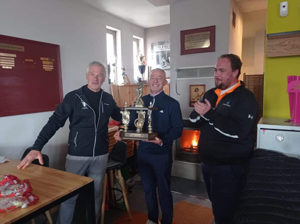 Célébration d'une victoire au golf avec des participants souriants tenant un trophée. L'image montre trois hommes dans un club de golf, avec un feu de cheminée en arrière-plan. Le gagnant, au centre, affiche une grande joie, tandis que les deux autres applaudissent. Des trophées sont visibles sur les étagères, soulignant l'esprit compétitif et convivial de l'événement. Cette scène illustre l'importance des compétitions de golf et des moments part