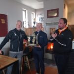 Célébration d'une victoire au golf avec des participants souriants tenant un trophée. L'image montre trois hommes dans un club de golf, avec un feu de cheminée en arrière-plan. Le gagnant, au centre, affiche une grande joie, tandis que les deux autres applaudissent. Des trophées sont visibles sur les étagères, soulignant l'esprit compétitif et convivial de l'événement. Cette scène illustre l'importance des compétitions de golf et des moments part