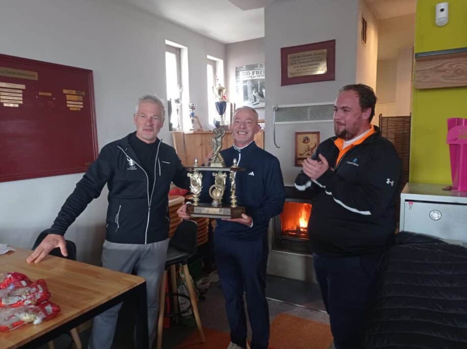 Célébration d'une victoire au golf avec des participants souriants tenant un trophée. L'image montre trois hommes dans un club de golf, avec un feu de cheminée en arrière-plan. Le gagnant, au centre, affiche une grande joie, tandis que les deux autres applaudissent. Des trophées sont visibles sur les étagères, soulignant l'esprit compétitif et convivial de l'événement. Cette scène illustre l'importance des compétitions de golf et des moments part