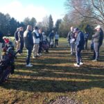 Groupe de golfeurs réunis sur un terrain de golf par une journée ensoleillée. Les participants, majoritairement des hommes, écoutent attentivement un instructeur qui donne des explications. Des sacs de golf sont visibles en arrière-plan, prêts à être utilisés. L'image illustre une séance d'entraînement ou un briefing avant une compétition de golf, mettant en avant l'esprit communautaire et l'engagement des passionnés de ce sport.