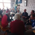 Groupe de personnes âgées célébrant un événement festif dans un club, portant des chapeaux de Noël. Ils sont assis autour d'une table, partageant des boissons et des collations. L'ambiance est conviviale et chaleureuse, illustrant des moments de joie et de camaraderie. En arrière-plan, on peut voir des éléments décoratifs liés au golf, évoquant un cadre de loisir et de rencontre. Cette image représente une belle occasion de socialisation et de partage
