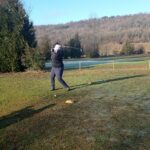 Un golfeur en train de frapper une balle sur un terrain de golf verdoyant, sous un ciel dégagé et lumineux. L'arrière-plan montre des collines boisées et des arbres, créant un cadre naturel paisible. Le golfeur porte un vêtement noir et un chapeau, concentré sur son swing. Des cônes de marquage sont visibles au sol, indiquant la zone de départ. Cette image illustre l'activité de golf en plein air,