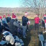 Groupe de golfeurs en tenue festive sur un parcours de golf, portant des bonnets de Noël. L'événement semble convivial, avec des participants attentifs à un discours donné par un homme au centre. On peut voir des chariots de golf à proximité, et le paysage environnant présente des arbres dénudés et un ciel clair. Cette scène évoque une activité de golf saisonnière, rassemblant des passionnés dans une ambiance joyeuse et festive.