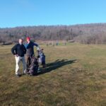 Deux golfeurs posent sur un terrain de golf verdoyant par une journée ensoleillée. L'un porte un pull noir et l'autre un gilet, tandis que des sacs de golf sont visibles à leurs côtés. En arrière-plan, plusieurs personnes jouent au golf sur un parcours entouré d'arbres dénudés, typique d'une saison hivernale. Cette image illustre une activité de loisirs en plein air, idéale pour les amateurs de golf.