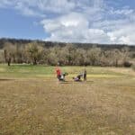 Image d'un terrain de golf au printemps, montrant des golfeurs en action. Un homme en t-shirt rouge se prépare à frapper sa balle, tandis qu'une femme avec une poussette de golf attend à proximité. En arrière-plan, des arbres dénudés et un ciel partiellement nuageux créent une atmosphère paisible. L'image illustre l'activité de golf en plein air, idéale pour les amateurs de ce sport.