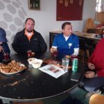 Quatre hommes se retrouvent autour d'une table dans un café, partageant un moment convivial. Ils dégustent des collations telles que des pâtisseries et des boissons, tandis qu'une assiette vide et des restes sont visibles sur la table. L'ambiance décontractée reflète un moment de camaraderie. En arrière-plan, on aperçoit des éléments décoratifs et des chaises empilées, témoignant d'un espace convivial. Ce rassemblement amical est idéal pour