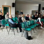 Salle de réunion avec un public attentif, composé d'une trentaine de personnes assises sur des chaises vertes. Les participants, d'âges variés, écoutent une présentation ou un discours. L'environnement est calme et bien éclairé, avec des murs clairs et un sol en carrelage. Des éléments de sécurité, comme un extincteur, sont visibles en arrière-plan, ajoutant à l'aspect formel de l'événement. Cette image illustre un