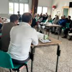 Réunion communautaire avec un groupe de participants attentifs, assis dans une salle. Au premier plan, plusieurs intervenants discutent autour d'une table, tandis qu'un public diversifié écoute. L'environnement est convivial, propice aux échanges et à la collaboration. Cette image illustre l'engagement civique et le dialogue au sein de la communauté.