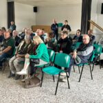Groupe de personnes assises dans une salle de réunion, écoutant attentivement une présentation. L'image montre une assemblée composée principalement de seniors, avec des chaises vertes disposées en rangées. L'environnement est lumineux et simple, propice à la discussion ou à un événement communautaire. Cette scène illustre l'engagement des membres de la communauté dans des activités sociales ou informatives.