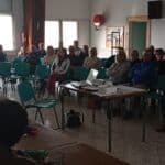 Réunion d'information avec un groupe de personnes assises dans une salle. L'audience, composée d'hommes et de femmes de divers âges, écoute attentivement un présentateur. Sur la table, un projecteur et des documents sont visibles, suggérant une présentation en cours. L'environnement est lumineux avec de grandes fenêtres et des chaises vertes. Cette scène illustre l'engagement communautaire et l'échange d'idées lors d'événements publics.