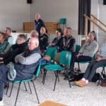 Groupe de personnes âgées réunies dans une salle pour assister à une présentation ou une conférence. Les participants, assis sur des chaises vertes, montrent un intérêt marqué pour le sujet abordé. L'environnement est lumineux et accueillant, favorisant l'échange et la convivialité. Cette image illustre l'engagement des seniors dans des activités sociales et culturelles.