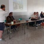 Réunion professionnelle en cours avec un orateur présentant des informations visuelles. Plusieurs participants, assis autour d'une table, écoutent attentivement. L'environnement est sobre, avec des chaises vertes et un écran projetant une image, probablement liée à un projet ou une initiative. Cette scène illustre l'importance de la collaboration et de la communication dans un cadre de travail.