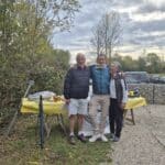 Trois personnes posent joyeusement autour d'une table extérieure recouverte d'une nappe jaune, lors d'un événement convivial en plein air. L'arrière-plan présente une nature verdoyante avec des arbres et un ciel nuageux. Sur la table, divers objets sont disposés, probablement liés à la célébration. L'atmosphère est festive, suggérant un moment de partage et de bonne humeur entre amis ou en famille.