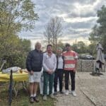Quatre personnes posent ensemble à l'extérieur, entourées d'une nature automnale, avec des arbres et un ciel nuageux en arrière-plan. Elles se tiennent près d'une table recouverte d'une nappe jaune, où sont disposés divers objets. L'ambiance décontractée suggère un événement convivial, probablement en lien avec une activité en plein air. Des voitures sont garées à l'arrière-plan, ajoutant une touche de vie à la scène.
