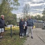 Quatre personnes se tiennent près d'une table en extérieur, entourées d'une végétation verdoyante. L'une des personnes, un homme aux cheveux blancs, sourit en tenant un document. Une femme à sa droite, vêtue d'une veste noire, regarde également vers l'objectif. Un homme en pull gris et une femme en chemise blanche se tiennent à côté d'eux, partageant un moment convivial. En arrière-plan, on aperçoit des voitures stationnées et un