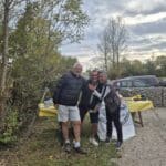 Trois personnes souriantes se tiennent devant une table recouverte d'une nappe jaune, dans un cadre naturel avec des arbres et un ciel nuageux en arrière-plan. L'ambiance est conviviale et décontractée, suggérant un événement en plein air, probablement lié à une activité sportive ou communautaire. Des accessoires de sport sont visibles sur la table, indiquant une connexion avec des activités de plein air.