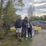 Trois personnes posent ensemble dans un cadre naturel, entourées d'arbres et de verdure. À l'arrière-plan, on aperçoit une table avec une nappe jaune, des assiettes et des ustensiles, suggérant un événement convivial en plein air. Le ciel est nuageux, indiquant une journée d'automne. Des voitures sont garées à proximité, ajoutant une touche de vie à la scène. Cette image évoque des moments de partage et