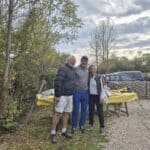 Trois personnes posent joyeusement ensemble en extérieur, entourées de verdure. À l'arrière-plan, on aperçoit une table avec des ustensiles, ainsi qu'une voiture stationnée. Le ciel est nuageux, suggérant une journée d'automne. Cette image évoque un moment convivial, probablement lors d'une rencontre amicale ou d'un événement en plein air.