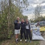 Trois personnes posent ensemble dans un cadre naturel, entourées d'arbres et de verdure. L'homme à gauche, portant un manteau noir, tient un document. Les deux femmes, vêtues de tenues sportives, sourient et affichent une ambiance amicale. En arrière-plan, on peut apercevoir une table avec des bouteilles de champagne et des objets de décoration, suggérant un événement festif. Cette image évoque un moment de célébration, probablement