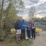 Quatre personnes souriantes se tiennent ensemble à l'extérieur, entourées par des arbres et un ciel nuageux. Elles posent devant une table jaune où sont disposés divers objets. L'atmosphère est conviviale, suggérant une célébration ou un événement communautaire. Cette image pourrait illustrer un rassemblement en plein air, une activité de groupe ou une remise de prix, mettant en avant l'esprit de camaraderie et de joie.