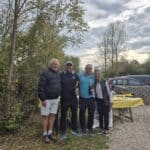 Quatre personnes posent ensemble à l'extérieur, entourées de verdure et d'un ciel nuageux. Deux hommes et deux femmes sourient tout en tenant des bouteilles, probablement en célébration. À l'arrière-plan, on peut apercevoir des voitures stationnées et une table avec des objets. Cette image évoque un moment convivial, peut-être lors d'un événement ou d'une fête en pleine nature.