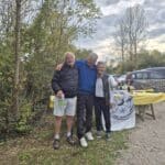 Trois personnes posent ensemble en extérieur, souriant et se tenant par les épaules, avec un décor de nature en arrière-plan. Un stand est visible à proximité, décoré d'une nappe jaune et portant un logo de golf, indiquant une activité ou un événement lié au golf. Des bouteilles de champagne et d'autres équipements sont disposés sur la table, suggérant une célébration ou un rassemblement convivial. L'atmosphère est chaleureuse et amicale, caractéristique