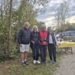 Groupe d'amis posant ensemble en plein air, entouré de verdure, lors d'un événement convivial. Ils portent des vêtements d'extérieur adaptés à la saison. En arrière-plan, on aperçoit une table avec une nappe jaune, probablement préparée pour un repas ou un apéritif, ainsi que des voitures stationnées. L'atmosphère décontractée et joyeuse reflète un moment de partage et de camaraderie.