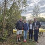 Quatre personnes posent ensemble en plein air, entourées d'une végétation verdoyante et d'un ciel nuageux. Elles se tiennent devant une table recouverte d'une nappe jaune, suggérant un événement convivial. L'atmosphère décontractée reflète un moment de partage et d'amitié, idéal pour des activités en groupe. Les participants portent des vêtements adaptés à la saison, indiquant une sortie automnale. Cette image évoque des rencontres sociales et des événements