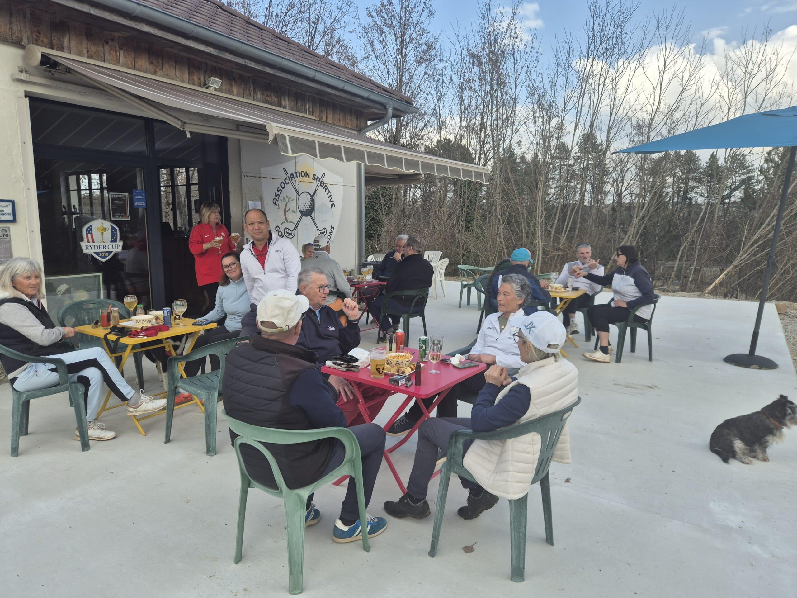 Groupe de personnes profitant d'un moment convivial sur la terrasse d'un café, entouré d'une nature verdoyante. Les clients, assis à des tables colorées, partagent des boissons et des en-cas tout en discutant. L'ambiance décontractée est accentuée par la présence d'un chien. En arrière-plan, un panneau indique l'association sportive, ajoutant une touche communautaire à l'événement. Ce cadre idéal invite à la détente et à l'é