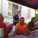 Groupe d'hommes réunis autour d'une table en extérieur, sous un abri, partageant un moment convivial après une activité. L'un des participants, en t-shirt orange, consulte son téléphone, tandis que d'autres discutent, sourient et savourent un repas. En arrière-plan, une maison avec des fenêtres ouvertes et un décor naturel. L'ambiance est détendue et amicale, reflétant un moment de camaraderie.
