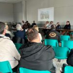 Réunion en cours dans une salle communautaire, avec des participants assis sur des chaises vertes. À l'avant, un groupe de personnes discute autour d'une table. Un écran projetant des informations est visible en arrière-plan. L'événement semble centré sur des sujets d'intérêt local, avec une atmosphère d'échange et de participation citoyenne.