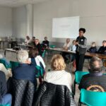 Réunion de la commission partenariat avec des participants attentifs dans une salle. Une intervenante se tient debout, présentant des informations sur un projet ou une initiative. À l'arrière-plan, un groupe de personnes, dont des membres du comité, écoute attentivement. Des équipements tels qu'un ordinateur et un projecteur sont visibles. L'événement semble axé sur le dialogue communautaire et le renforcement des partenariats locaux.