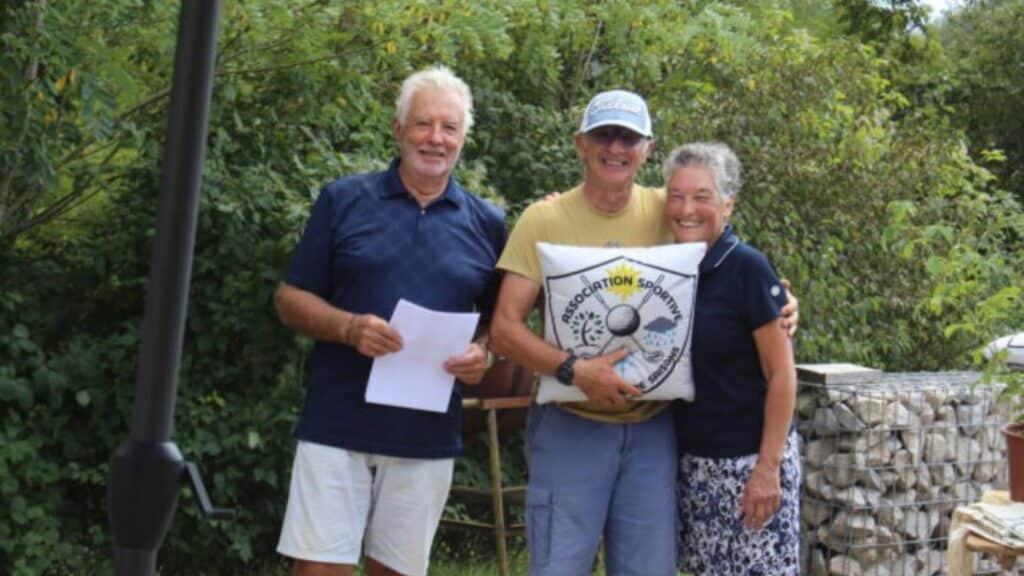 Trois personnes posent ensemble à l'extérieur, entourées de verdure. L'homme au centre tient un coussin avec le logo d'une association sportive. Les deux autres, un homme à gauche et une femme à droite, sourient et semblent partager un moment convivial. En arrière-plan, on aperçoit des murets en pierre et des éléments de jardinage, suggérant une ambiance chaleureuse et amicale. Cette image évoque des activités communautaires et un engagement envers le sport.