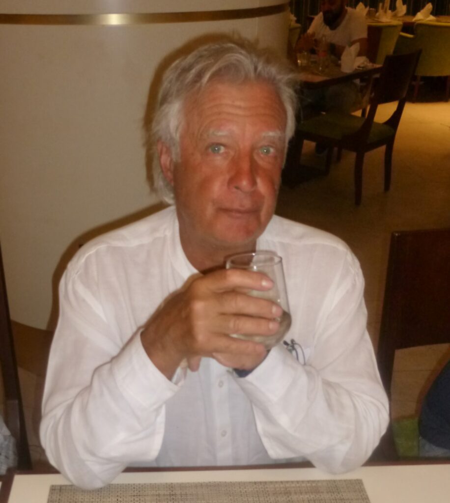 Homme aux cheveux gris, portant une chemise blanche, assis à une table dans un restaurant. Il tient un verre transparent dans ses mains, affichant une expression détendue. En arrière-plan, des convives sont visibles, créant une ambiance conviviale. Cette image illustre une scène de détente et de convivialité dans un cadre gastronomique.