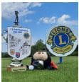 Image représentant un événement sportif local avec un trophée en premier plan, un personnage en peluche aux couleurs de l'équipe, et des panneaux signalant le club Lions et le championnat. Le ciel est dégagé, et l'environnement est verdoyant, symbolisant une journée festive dédiée au sport et à la communauté.