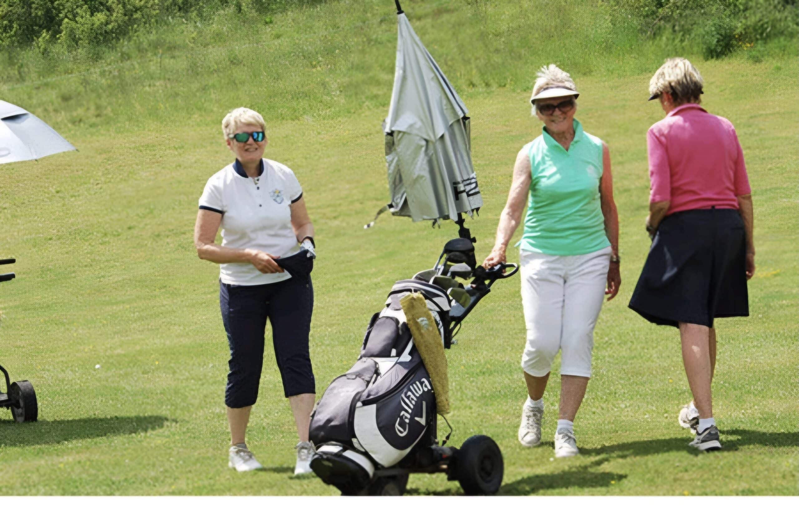 Trois femmes jouent au golf sur un parcours verdoyant. L'une d'elles, portant un polo blanc et des lunettes de soleil, sourit tout en tenant un chapeau. Une autre femme, vêtue d'un débardeur vert et de pantalons blancs, pousse une voiture de golf équipée d'un parapluie. La troisième femme, en haut rose, se dirige vers elles. Cette scène évoque une journée ensoleillée de golf, idéale pour les amateurs du sport.