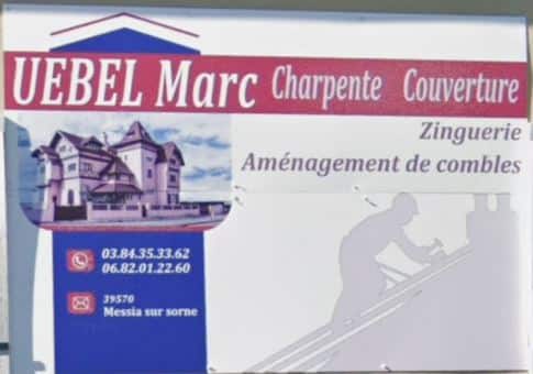 Panneau d'affichage de l'entreprise UEBEL Marc, spécialisée en charpente et couverture. Le visuel présente une maison au style architectural distinctif. Les services proposés incluent la zinguerie et l'aménagement de combles. Coordonnées de contact : 03.84.35.33.62 et 06.82.01.22.60, localisée à Messia sur Sorne.
