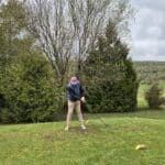Un golfeur se prépare à frapper une balle sur un fairway verdoyant, entouré d'arbres feuillus. Il porte un chapeau rose et un gilet bleu, adoptant une posture concentrée pour son swing. Au sol, des repères jaunes indiquent l'emplacement des tees. Le ciel est nuageux, créant une ambiance typique d'une journée de golf. Cette image évoque la passion du golf et l'importance de la technique dans ce