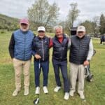 Quatre golfeurs posent ensemble sur un terrain de golf verdoyant. Ils portent des vestes sans manches et des pantalons confortables, prêts à profiter d'une journée de golf. L'arrière-plan présente un ciel nuageux et des arbres, créant une atmosphère paisible. Les golfeurs affichent des sourires amicaux, soulignant l'esprit de camaraderie et de passion pour le golf. Cette image évoque les plaisirs du sport en plein air et